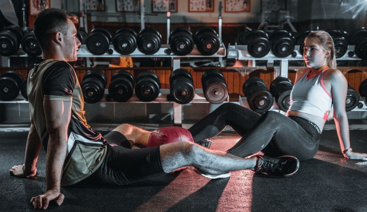 A man and woman resting together on the gym floor, surrounded by dumbbells.