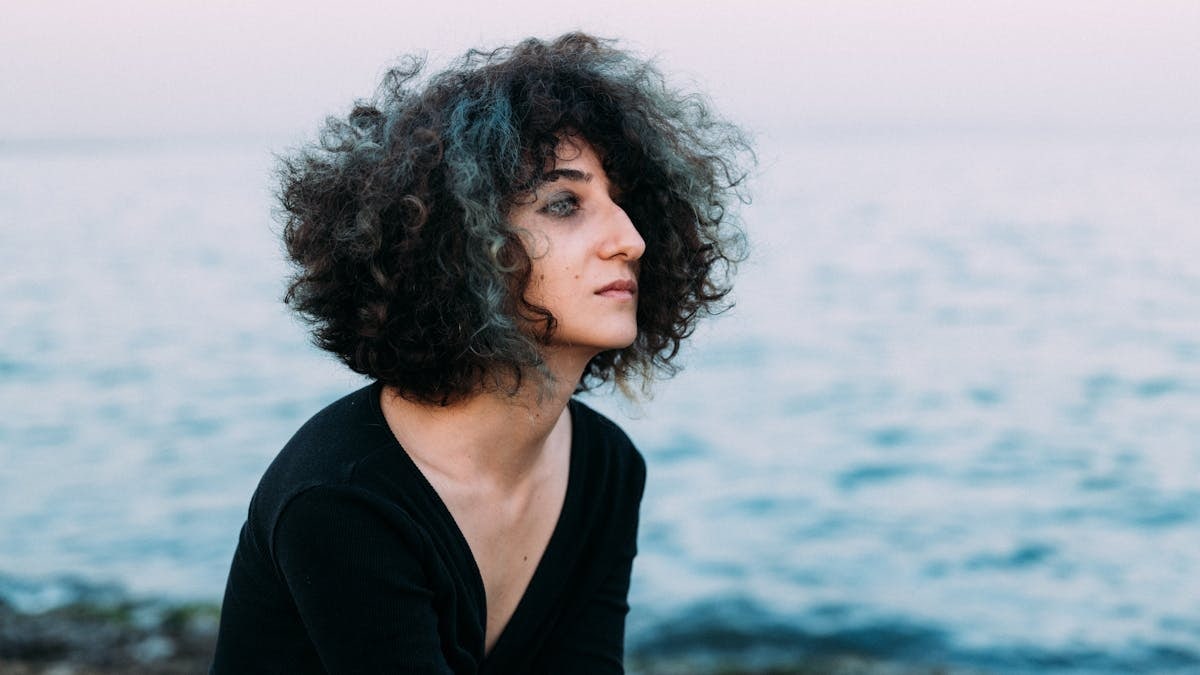 A woman with curly hair gazes at the horizon by the sea during sunset.